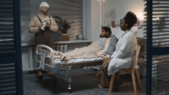 Vídeo curto decorativo animado, sem som e repetitivo, de um palhaço branco engraçado, brincando com uma menina preta pequena deitada na cama do hospital, entretendo-a e fazendo-a rir, com uma jovem médica preta amigável sentada por perto.