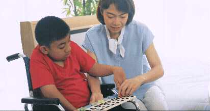 Vídeo curto decorativo animado, sem som e repetitivo, de um menino amarelo sentado em uma cadeira de rodas, segurando um quadro de encaixe com números em alto relevo, sendo ajudado e ensinado por uma professora amarela ao lado.