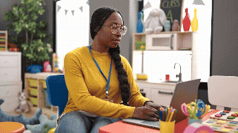 Vídeo curto decorativo animado, sem som e repetitivo, de uma professora preta, que usa óculos e tem longas tranças, que está em uma sala de aula para crianças, sentada e escrevendo em um notebook. Ela olha para a câmera e sorri.