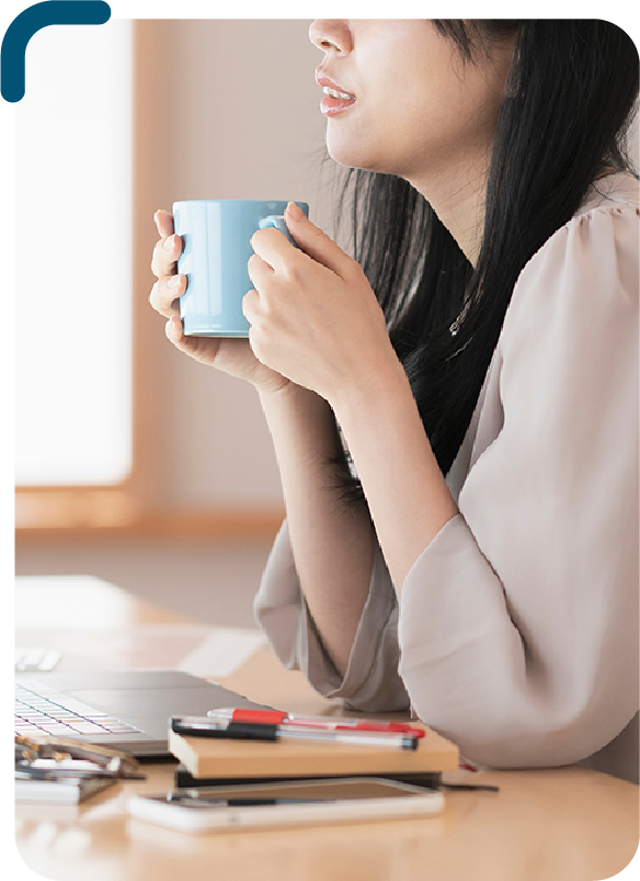 foto de uma mulher branca sentada na frente de uma mesa, em cima da mesa tem um laptop aberto e algumas coisas como caneta e agenda ao lado. A mulher está vestindo uma blusa cinza e está segurando uma caneca azul com as duas mãos, apoiando os cotovelos na mesa.