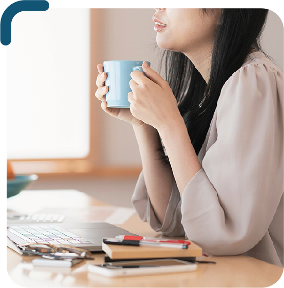 foto de uma mulher branca sentada na frente de uma mesa, em cima da mesa tem um laptop aberto e algumas coisas como caneta e agenda ao lado. A mulher está vestindo uma blusa cinza e está segurando uma caneca azul com as duas mãos, apoiando os cotovelos na mesa.