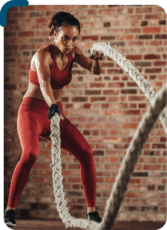 Foto colorida de uma mulher negra de pele clara, com roupa de ginástica, segurando uma corda em cada mão, fazendo movimentos para cima e para baixo, intercalando as mãos. Ao fundo tem uma parede de tijolos aparentes.