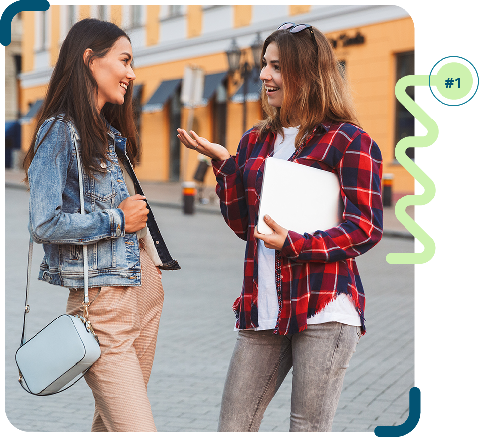 Foto colorida de duas mulheres de pele clara, conversando na rua de paralelepípedo. Uma está vestindo calça clara, jaqueta jeans e bolsa azul; a outra está de calça jeans, camiseta branca e camisa xadrez vermelha e preta. 