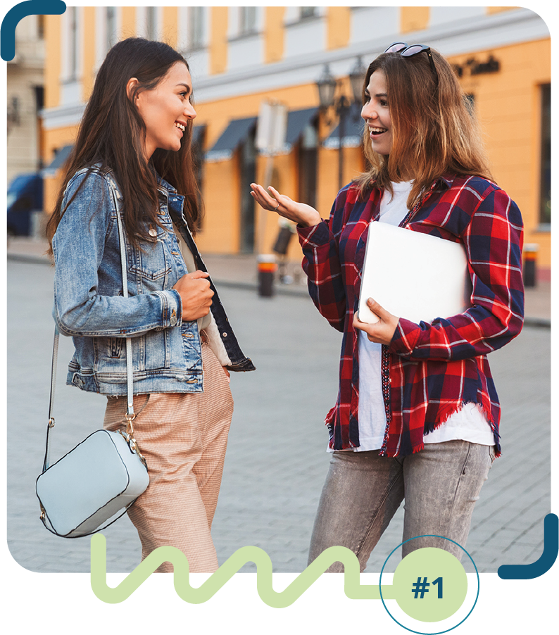 Foto colorida de duas mulheres de pele clara, conversando na rua de paralelepípedo. Uma está vestindo calça clara, jaqueta jeans e bolsa azul; a outra está de calça jeans, camiseta branca e camisa xadrez vermelha e preta. 