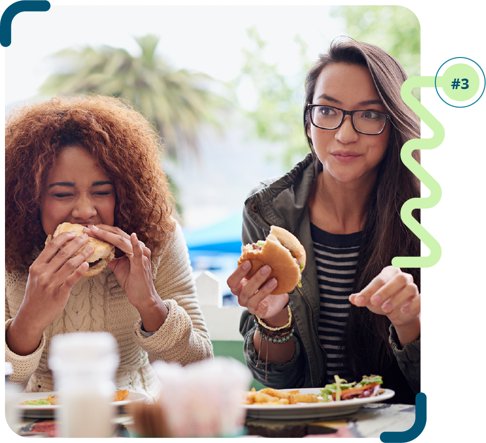 Foto colorida de duas mulheres (uma negra e uma de pele clara) sentadas em frente a uma mesa, comendo sanduíche, uma está mordendo o sanduíche e a outra está mastigando.