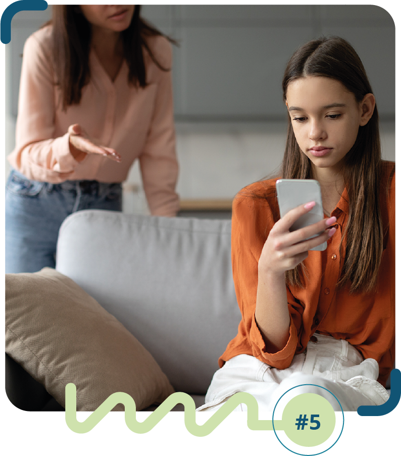 Foto colorida de uma adolescente sentada em um sofá, mexendo em um celular e uma adulta em pé ao lado do sofá falando com a adolescente.