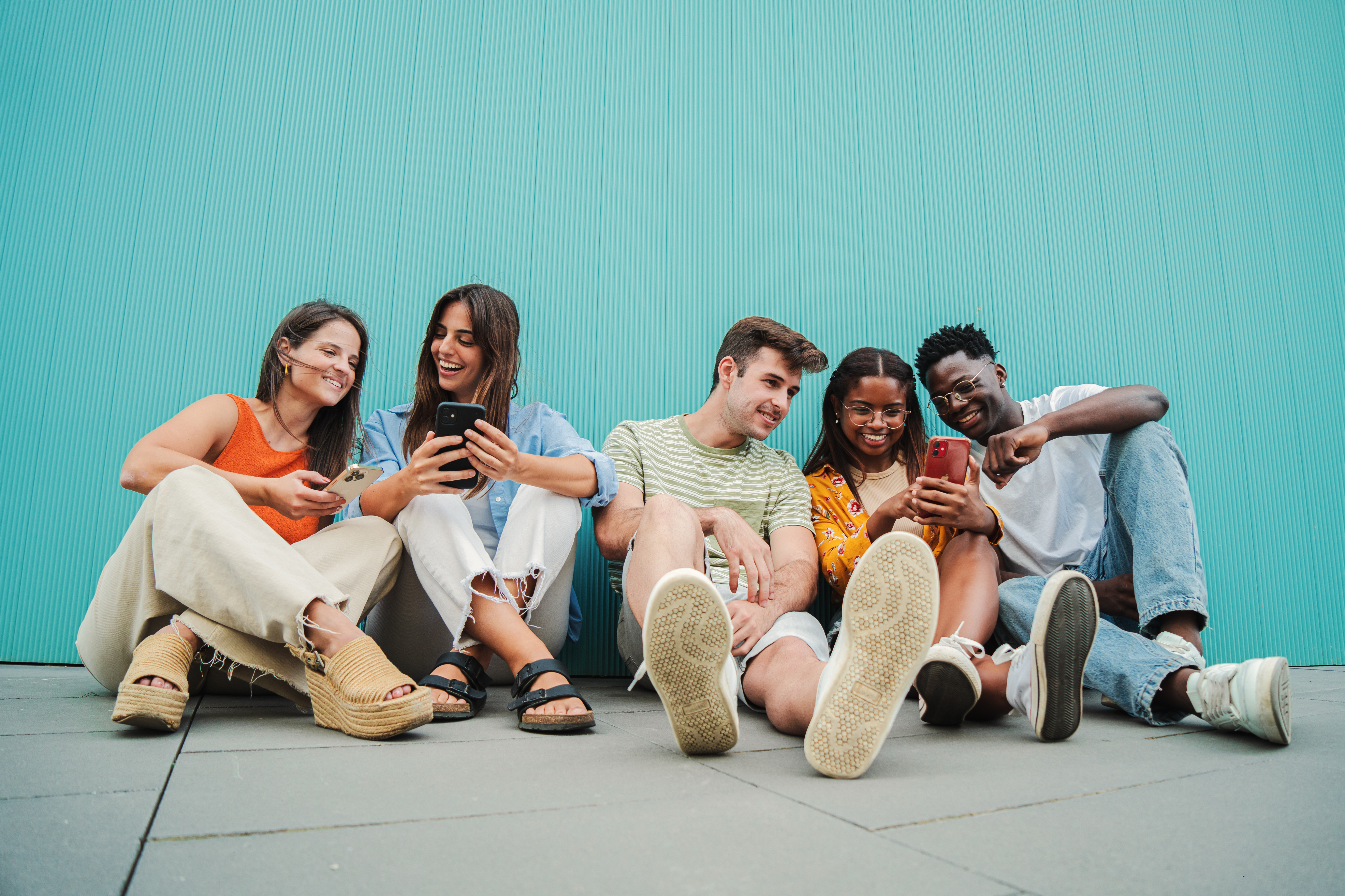 Grupo de 5 pessoas sentadas no chão e encostadas em uma parede azul. Da esquerda para direita tem: 1 - mulher branca, vestida com uma regata laranja, uma calça e uma sandália bege, ela está sorrindo, com um celular na não e está olhando para o celular da pessoa dois. 2 - mulher branca, vestindo uma camisa e uma calça jeans, e uma sandália preta, está segurando um celular e interagindo com a pessoa um. 3 - homem branco vestindo uma camisa verde listrada, short jeans e tênis, está olhando para o celular da quarta pessoa. 4 - mulher negra, vestindo camiseta bege, uma camisa laranja e um tênis branco, está com um celular na mão e interagindo com as pessoas 3 e 5. 5 - homem negro, vestindo uma camiseta azul clara, calça jeans e tênis branco, está interagindo com a pessoa 4.