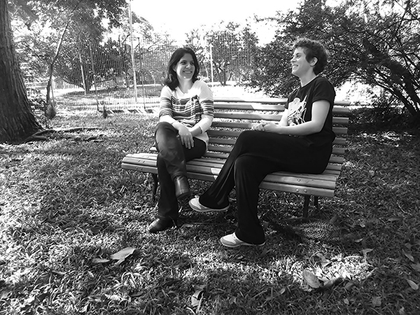 Duas mulheres conversam sentadas em um banco, em local aberto. A fotografia mostra ambas de corpo inteiro.