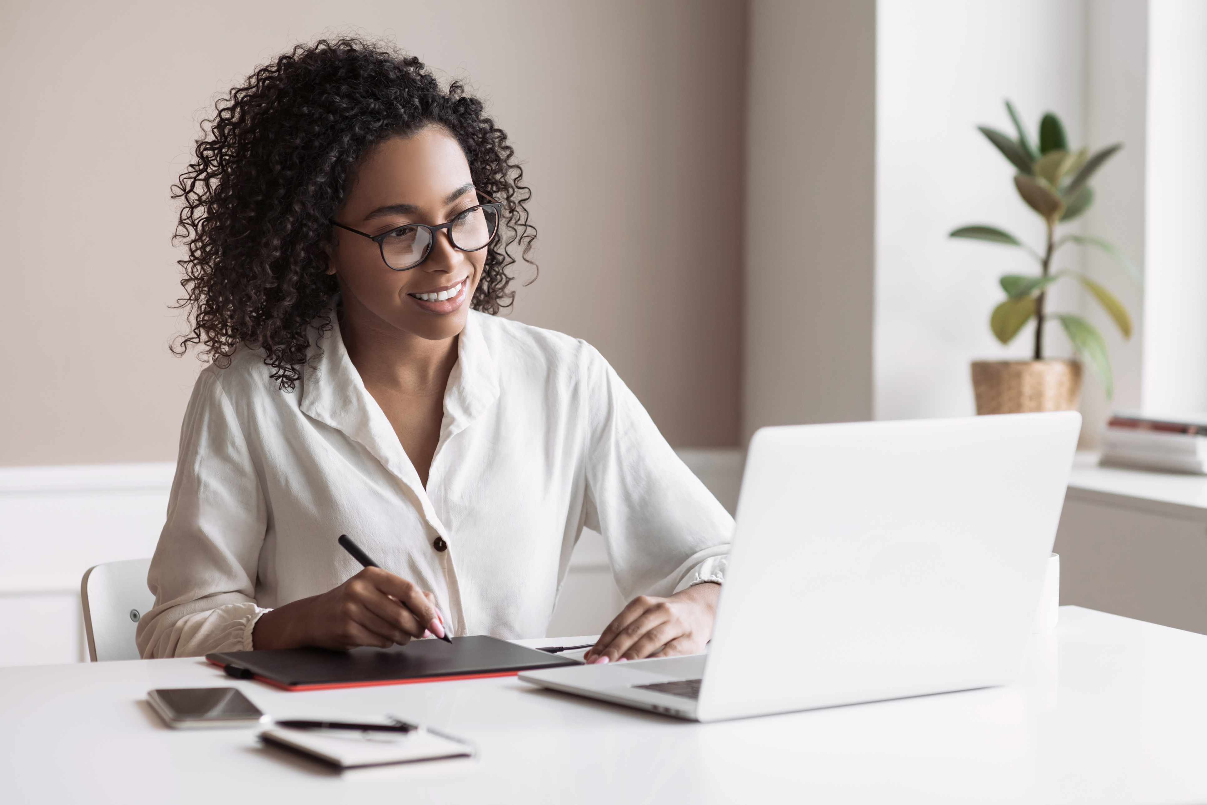 Mulher falando ao telefone sorrindo, sentada diante de uma mesa onde tem alguns papéis e um notebook. Ela olha para os papéis.