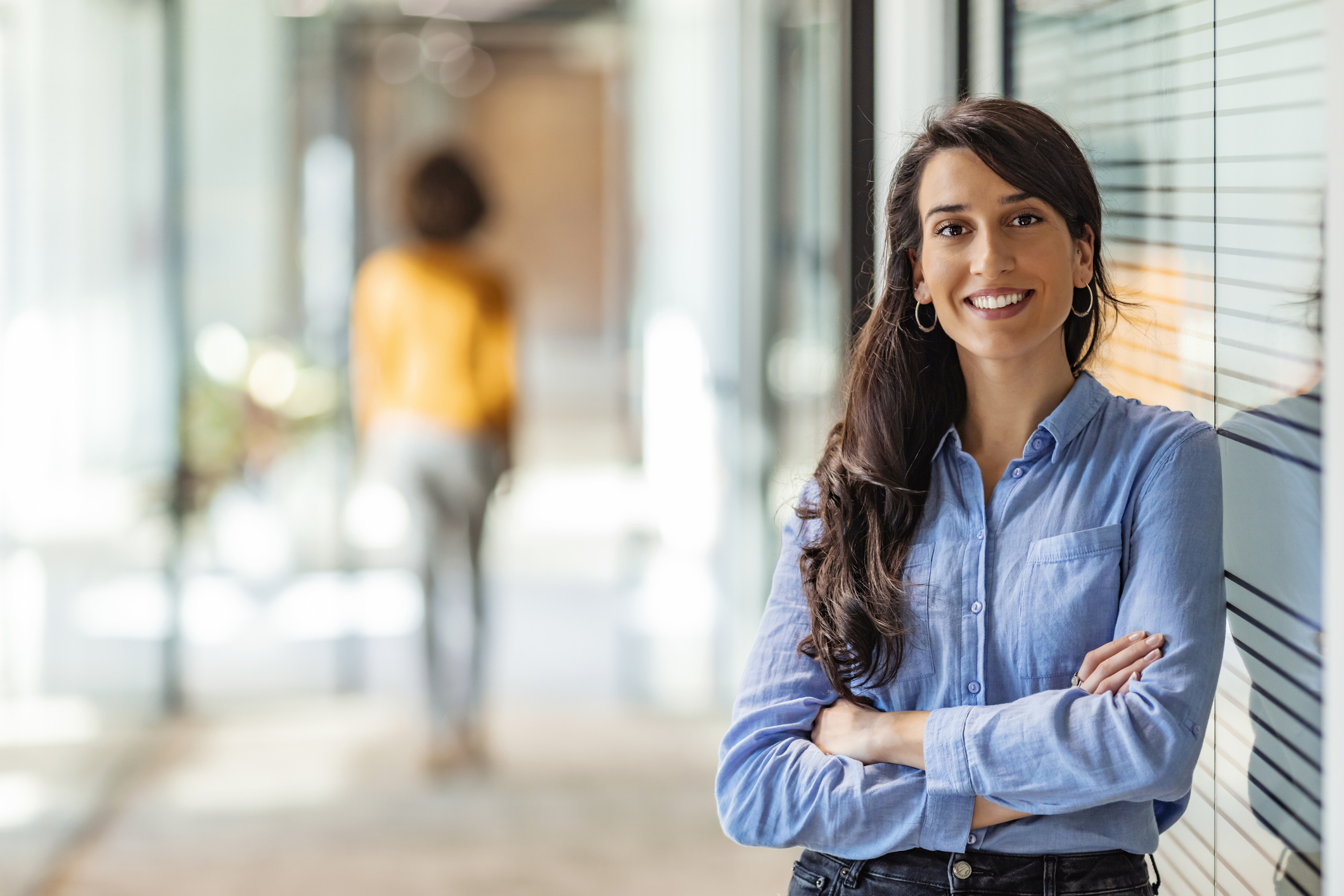 A imagem mostra uma mulher sorrindo e de braços cruzados em seu corredor de trabalho.